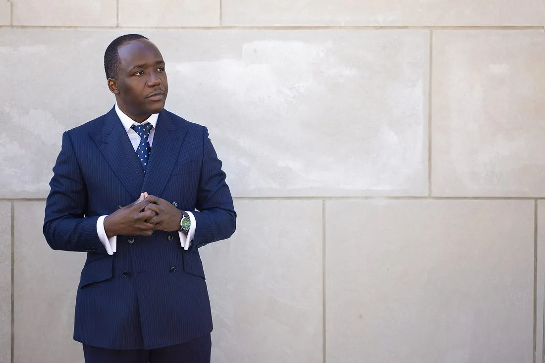Dr. Job Mogire in a navy pinstripe suit against a limestone wall, gaze off camera